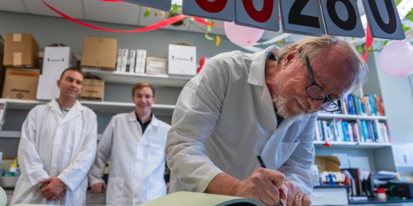 Richard Lenski updates the lab book for the Long Term Evolution Experiment.  Lab partner Jeff Barrick (left) and lab manager Devin Lake (right) look on from the background. Photo:  Finn Gomez